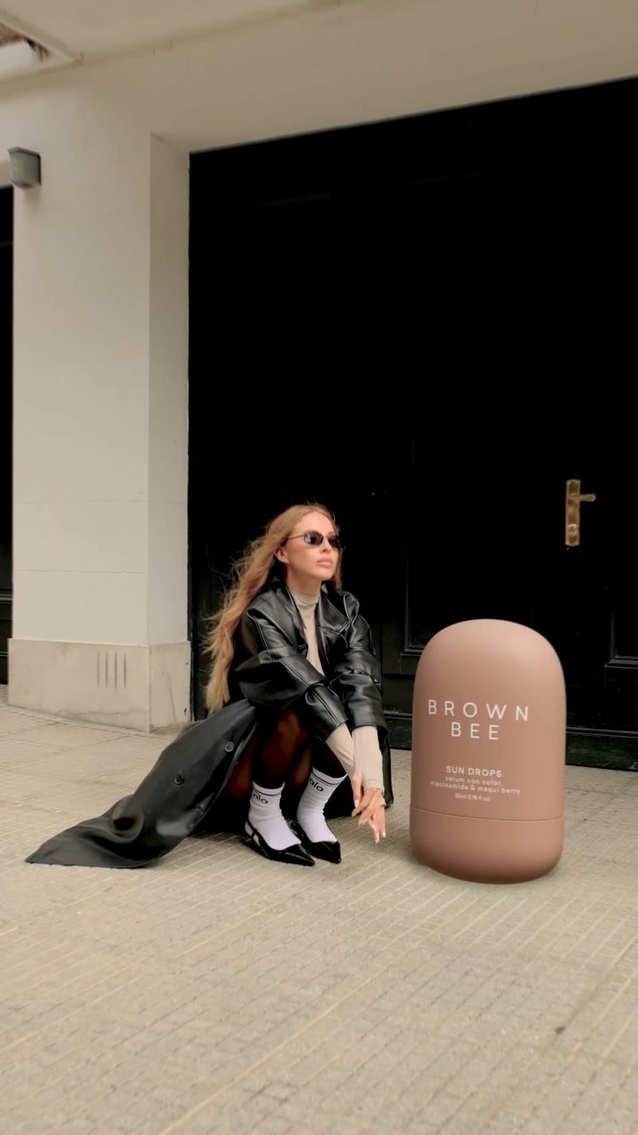 Una mujer con abrigo negro se sienta junto a un envase gigante de Sun Drops de Brown Bee en una vereda