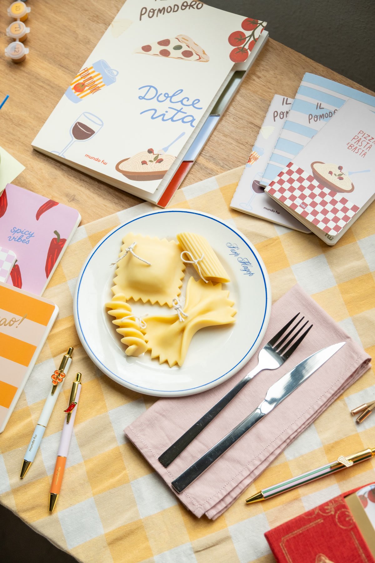 Cuadernos Dolce Vita de FW y velas con forma de pasta sobre una mesa con mantel a cuadros