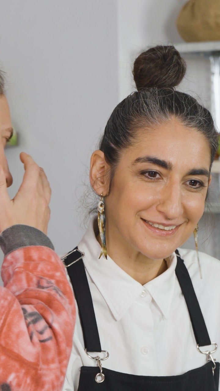 Una mujer con camisa blanca y delantal negro sonríe durante una escena de cocina